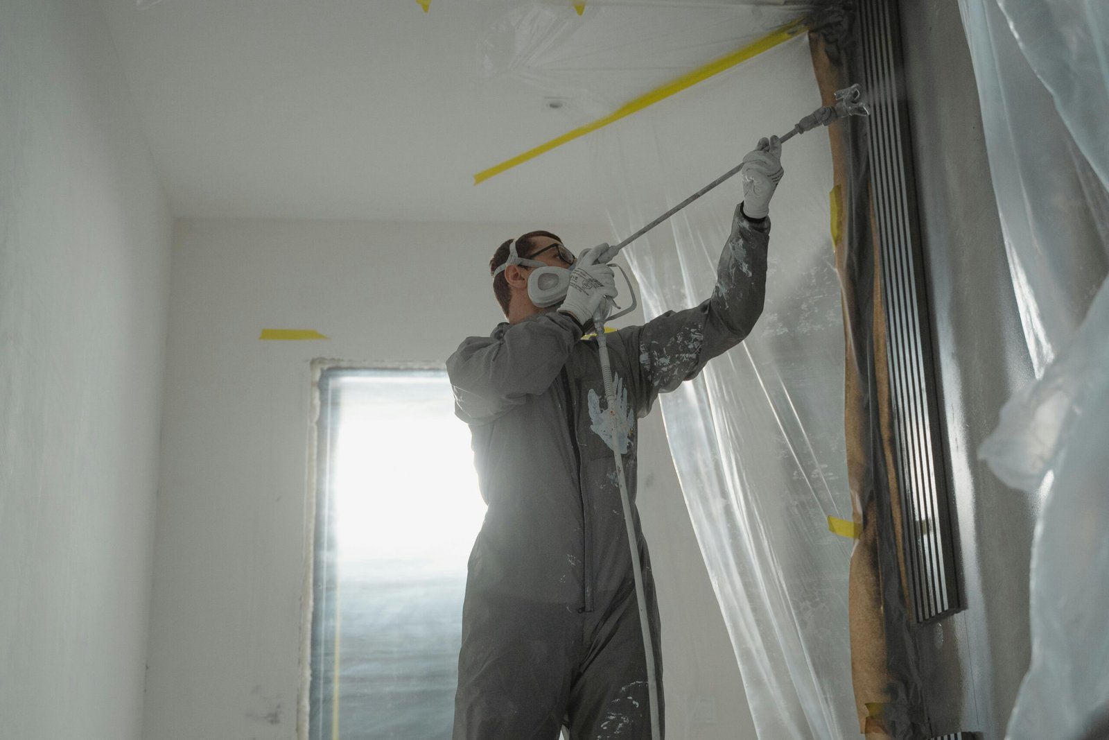 A professional painter wearing protective gear spray paints a wall indoors, working on a home renovation project.